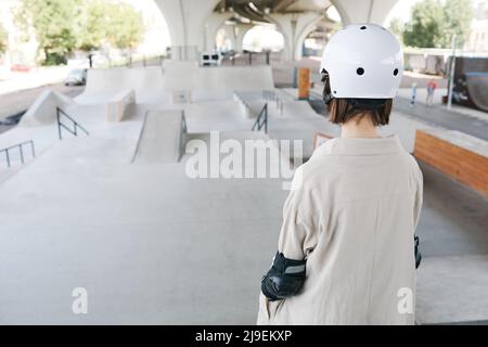 Back view of young teenage girl wearing protective guards and helmet in skating park outdoors, copy space Stock Photo
