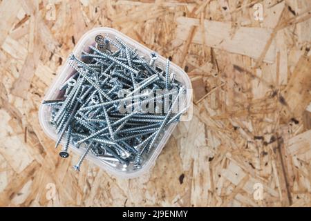 Shiny new self-tapping screws are in plastic box standing on a wooden floor, top view. Close-up background photo with selective soft focus Stock Photo