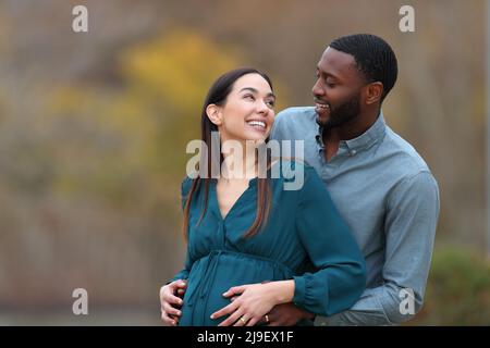 Interracial couple looking each other enjoying pregnancy in a park Stock Photo
