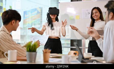 A group of cheerful millennial business startup having a meeting in the office, trying a new innovation, wearing a VR headset. Stock Photo
