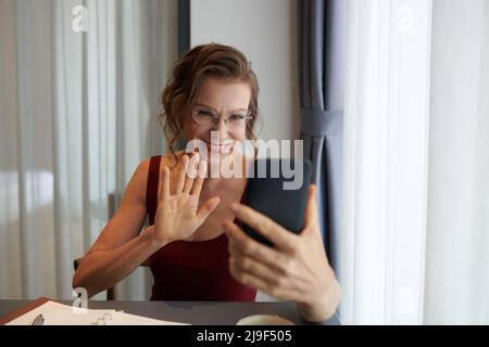 Positive elelegant businesswoman in glasses waving with hand when video calling her coworker or family memeber Stock Photo