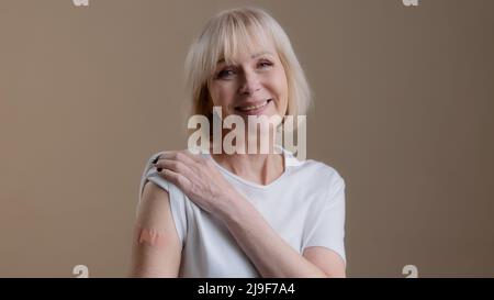 Coronavirus immunization infection covid19 vaccination happy vaccinated senior lady mature woman posing with adhesive medical plaster on arm after Stock Photo