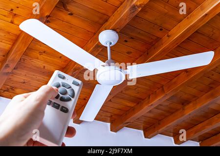 A man uses a remote control to turn on a white ceiling fan mounted in a house with wooden ceilings. Stock Photo