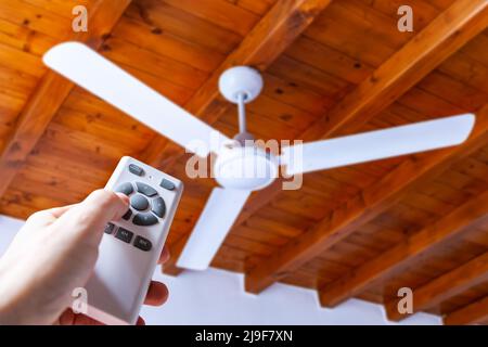 Close up shot of a hand using a remote control to operate a ceiling fan mounted in a house on a wooden ceiling. Stock Photo