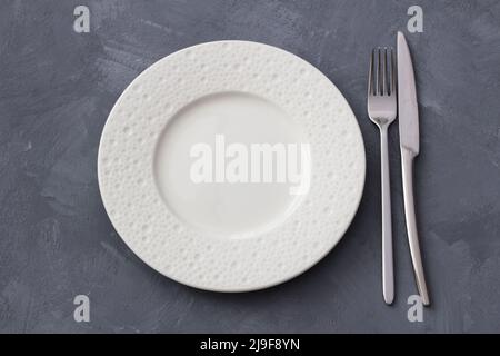 White ceramic plate on a gray textured background with fork and knife, top view, horizontal Stock Photo