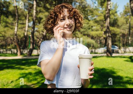 Young redhead woman wearing white tshirt holding takeaway coffee cup, talking with friends on mobile phone while standing on green city park, outdoors Stock Photo