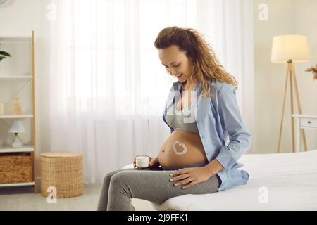 Pregnant woman in ninth month sits at home on bed and smears abdomen with anti-stretch mark cream. Stock Photo