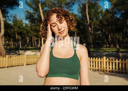 Young redhead woman wearing sportive clothes standing on city park suffering from headache desperate and stressed because pain and migraine. Hands on Stock Photo