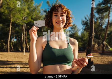 Young happy redhead woman wearing sports bra on city park, outdoors showing plastic credit or debit card while holding mobile phone. Online shopping, Stock Photo