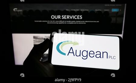 Person holding mobile phone with logo of British waste management company Augean plc on screen in front of web page. Focus on phone display. Stock Photo