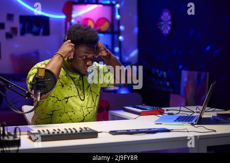 Portrait of young African American man wearing stylish neon T-shirt putting on headphones to start working on music track Stock Photo