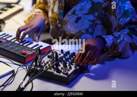 Unrecognizable young Black female musician sitting at desk working on music track adjusting sound settings using mixing console and synthesizer Stock Photo