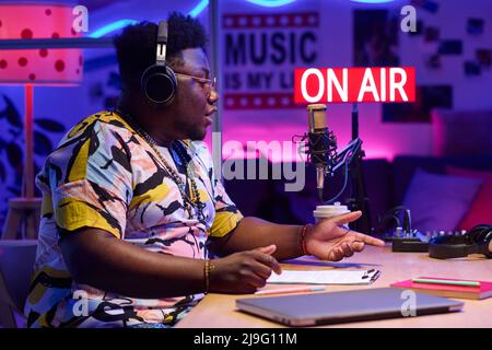 Side view of handsome young African American guy wearing headphones sitting at desk recording podcast or broadcast in home studio Stock Photo