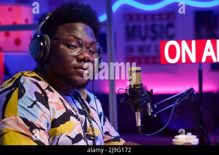 Medium close-up of self-confident young gen Z African American man wearing headphones doing broadcasting looking at camera Stock Photo