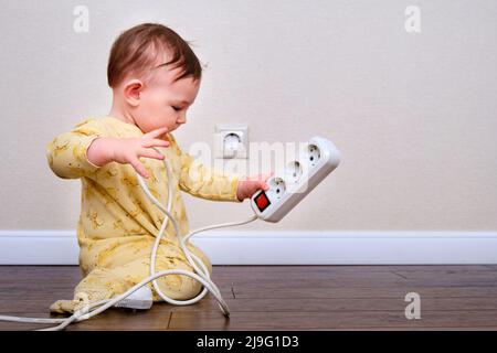 Toddler baby boy plays with electric wires while sitting on the floor. Child holding an extension cord with electrical sockets, copy space Stock Photo