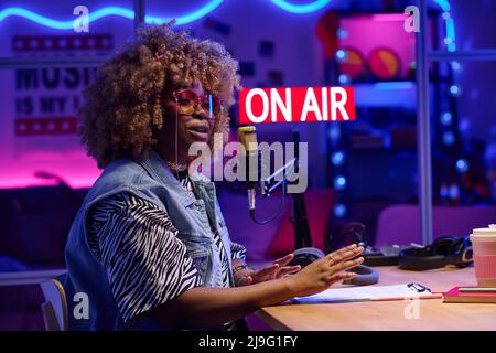 Portrait of stylish African American woman sitting at desk in podcasting or broadcasting studio speaking on topic Stock Photo