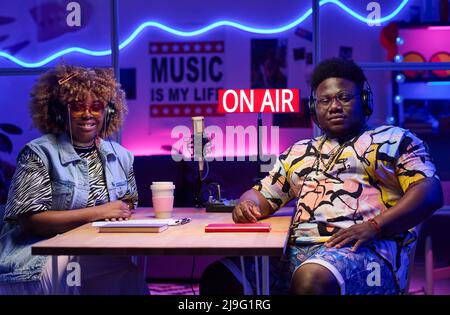 Young African American man and woman wearing fashionable outfits sitting at desk looking at camera after working together on podcast Stock Photo