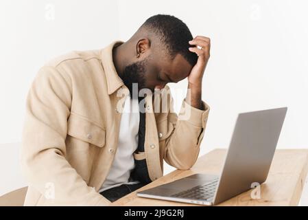 Tired depressed african american freelancer, frustrated with business failure, working at laptop, feeling exhausted with headache, upset at home. Stock Photo