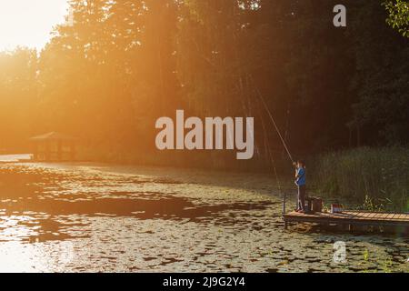 Little boy standing and fishing on a wooden dock at the sunset. Stock Photo