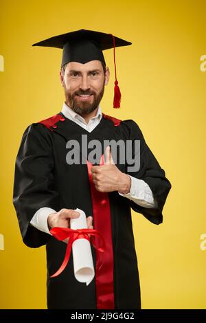 Front view of cheerful, proud student holding diploma, showing super. Handsome boy in mortarboard and graduate gown, smiling, looking at camera. Isolated on yellow backgrround. Stock Photo