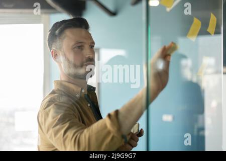 Male Entrepreneur Putting Sticky Notes On Glass Wall In Office Stock Photo