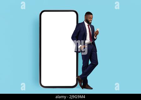 Business App. Black Businessman In Suit Standing Next To Big Blank Cellphone Stock Photo