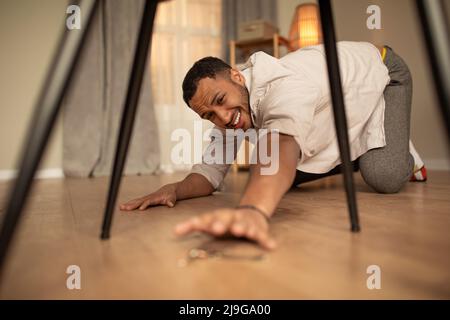 Handsome young black man standing on his knees, trying to reach house keys, dropping them on floor at home Stock Photo