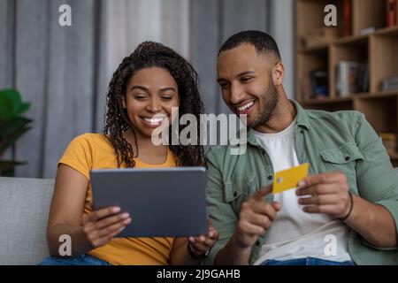 Happy black spouses shopping online using tablet and credit card, paying for order, sitting on sofa at home Stock Photo