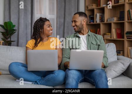 Young black couple using laptop computers, looking and smiling to each other, sitting on couch and surfing internet Stock Photo