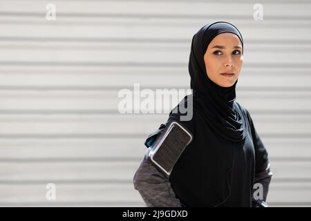 Confident serious calm pretty young muslim woman in hijab with phone on her shoulder resting from exercises Stock Photo