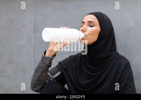Tired pretty young middle eastern woman in hijab drinks water from bottle on gray wall background outdoor Stock Photo