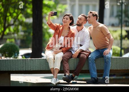 Smiling woman taking selfie with her friends or coworkers when sitting on park bench Stock Photo