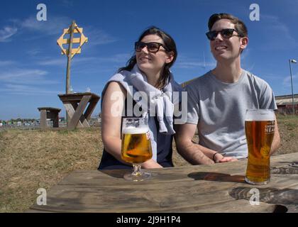 Young couple enjoying drinks on a sunny pub bench, looking into the distance. Bright, relaxed lifestyle stock photography of a male and female Stock Photo