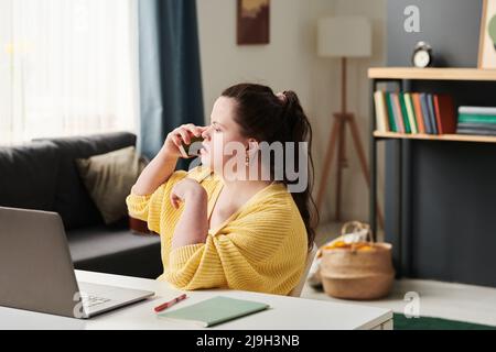 Young woman with Down syndrome using sitting at table with laptop on it talking on phone while working or studying remotely from home Stock Photo