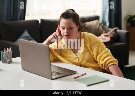 Young Woman with Down syndrome sitting relaxed at table at home with laptop on it talking on phone with friend or co-worker Stock Photo