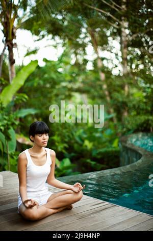 A young Asian woman meditates in a private pool villa at Kayumanis Ubud, Bali, Indonesia. Stock Photo