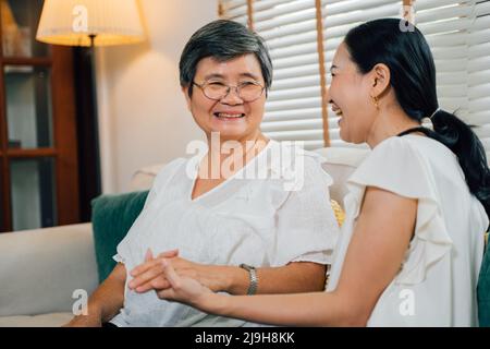 Cheerful elderly Asian mom and adult daughter holding hands and looking at each other while relaxing on sofa and laughing near window at home. Stock Photo