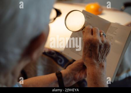 Senior woman with magnifying glass reading book at home Stock Photo