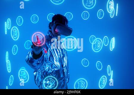 Woman wearing virtual reality simulator touching bitcoin symbol on screen against blue background Stock Photo