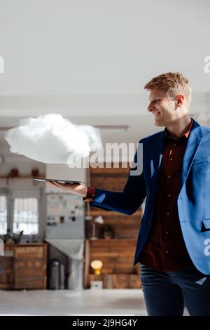 Smiling businessman holding tablet PC under levitating cloud network in office Stock Photo