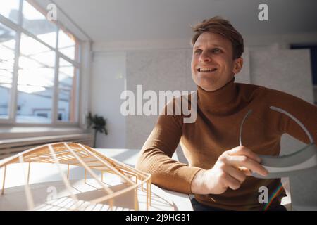 Smiling architect holding virtual reality simulator sitting by leaf shaped model at desk in office Stock Photo