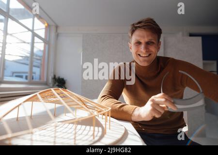 Happy architect holding virtual reality simulator sitting by leaf shaped model at desk in office Stock Photo