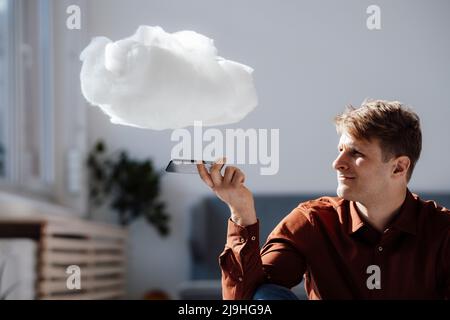 Businessman holding mobile phone under levitating cloud network in office Stock Photo