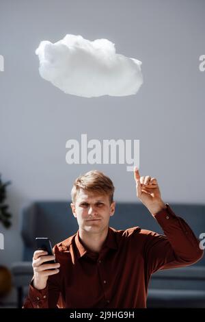 Smiling businessman holding mobile phone pointing at levitating cloud network in office Stock Photo