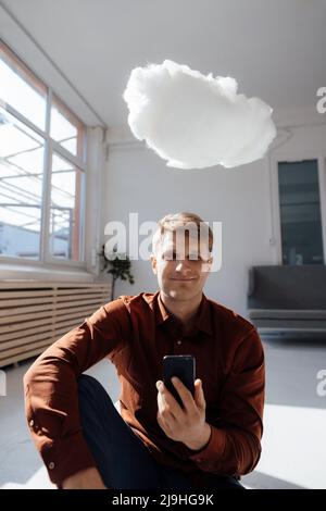 Smiling businessman holding smart phone sitting under levitating cloud network in office Stock Photo