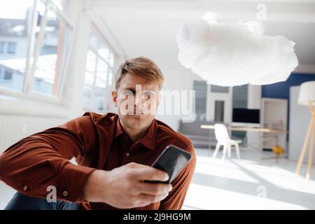 Businessman holding smart phone sitting by levitating cloud network in office Stock Photo