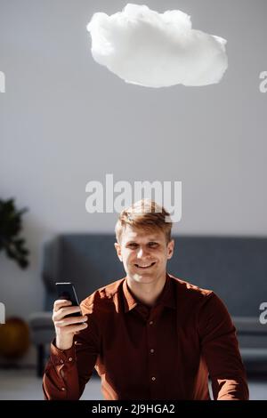 Smiling businessman holding mobile phone sitting under levitating cloud network in office Stock Photo
