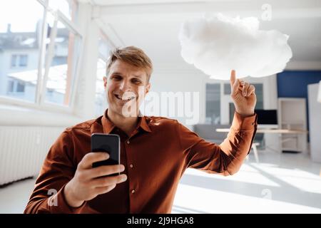 Happy businessman holding mobile phone pointing at levitating cloud network in office Stock Photo