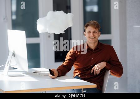 Happy businessman holding mobile phone sitting by levitating cloud network in office Stock Photo