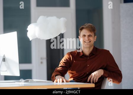 Smiling businessman with desktop PC sitting by levitating cloud network in office Stock Photo